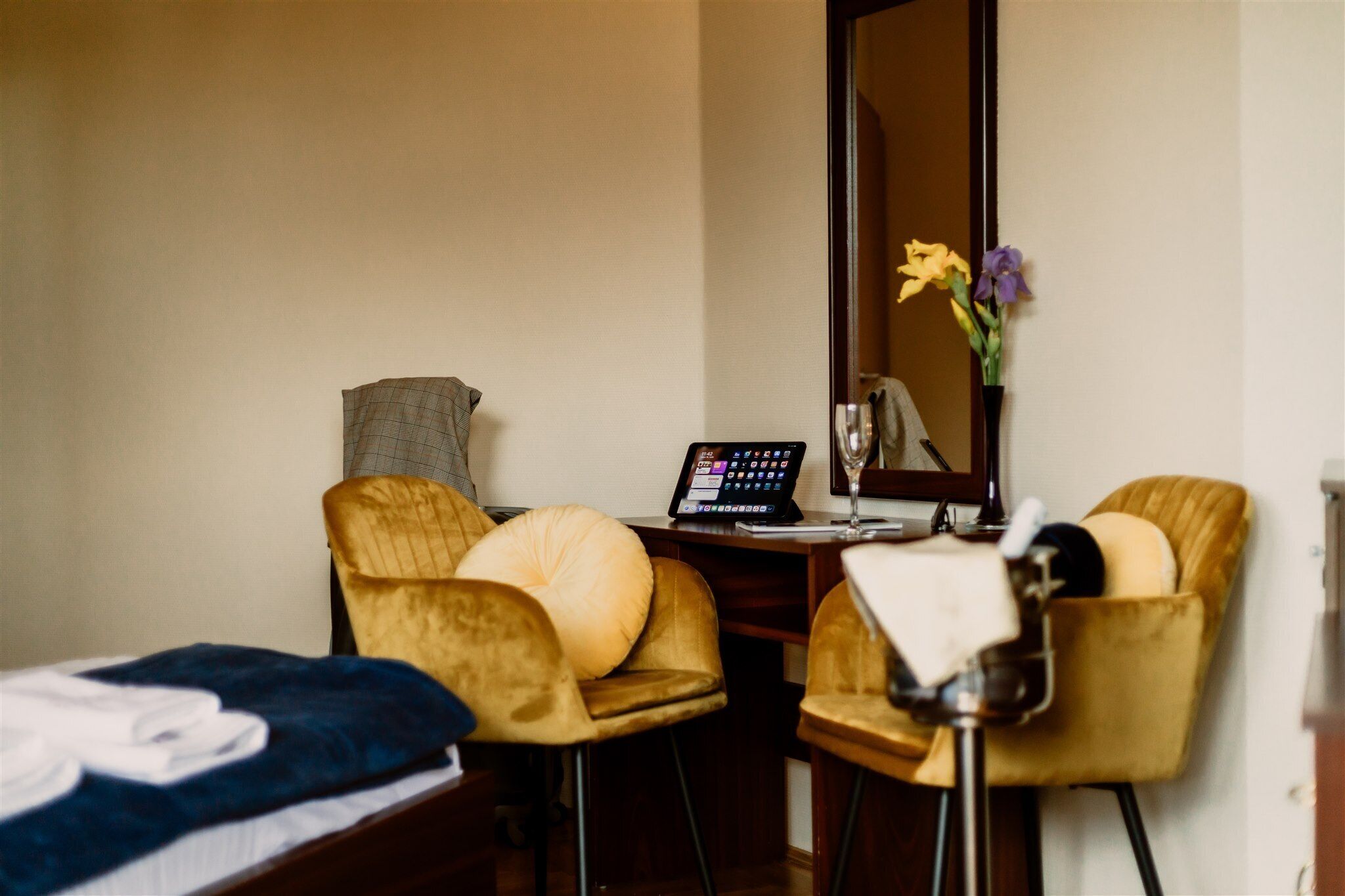 Habitación de hotel con cama, dos sillas, escritorio con una computadora portátil, espejo y un florero con flores.