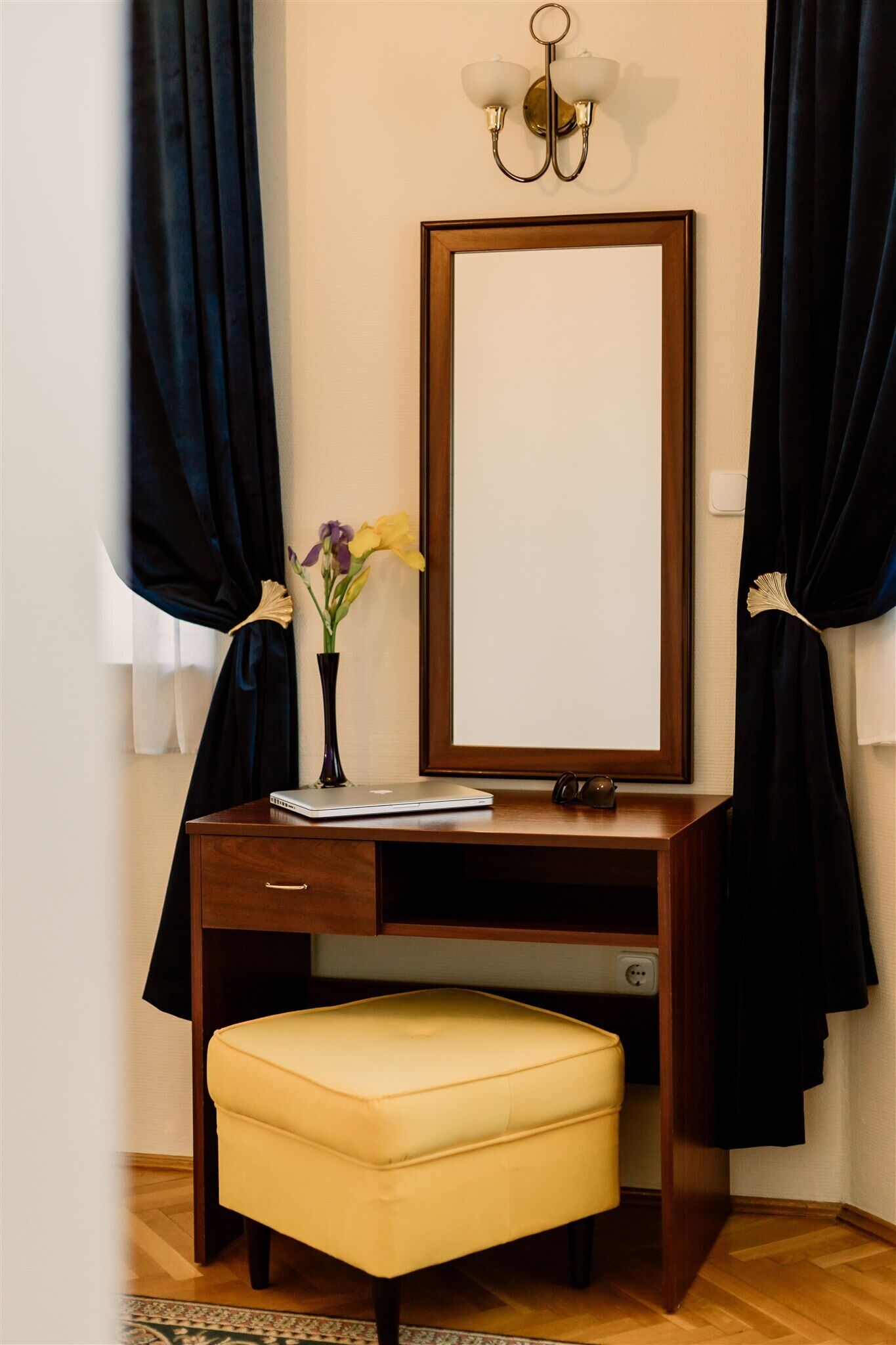 A wooden desk with a mirror, a laptop, and a vase with flowers.