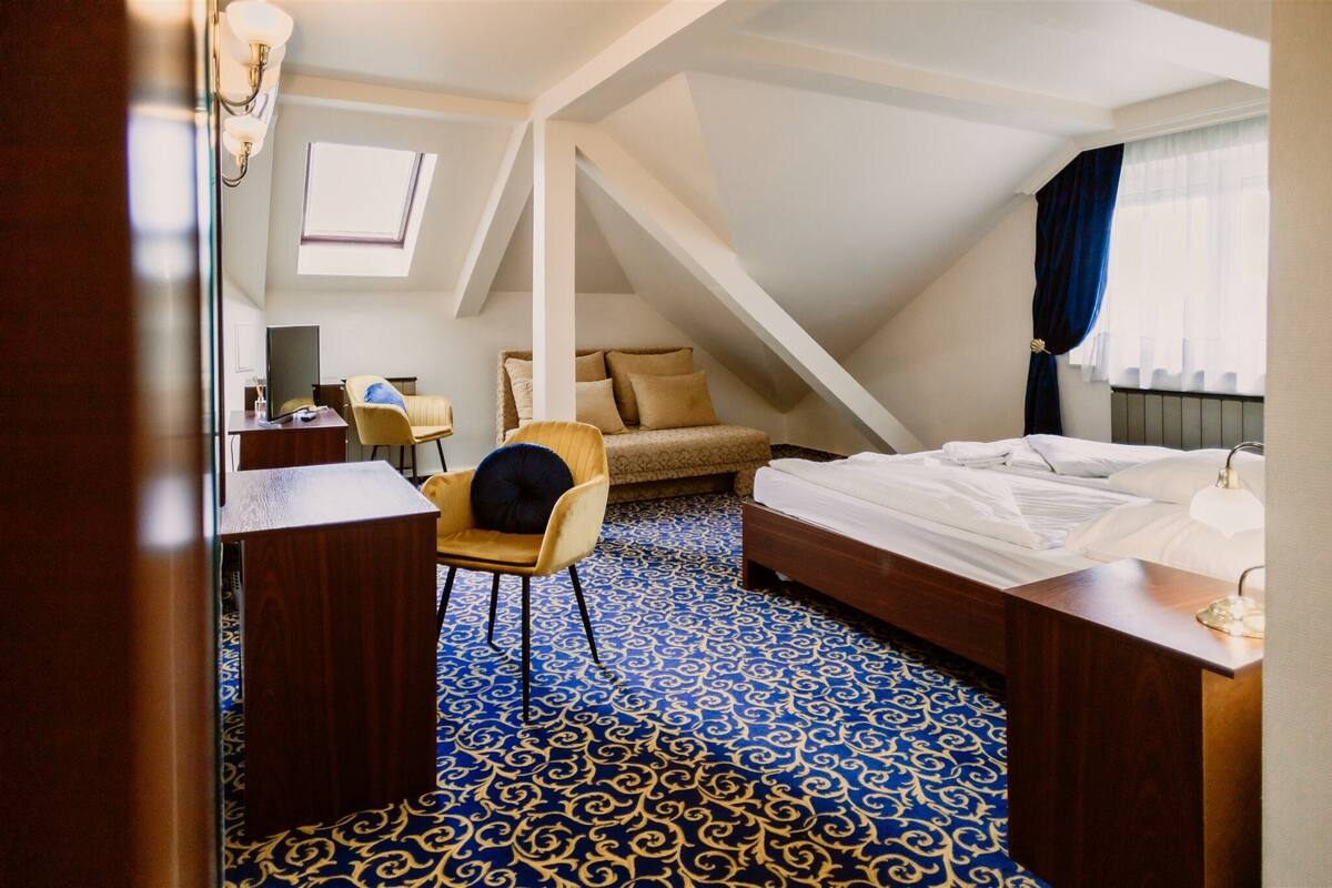 A hotel room with a sloped ceiling, a bed with white linens, a desk with a chair, a sofa with cushions, and a patterned carpet.