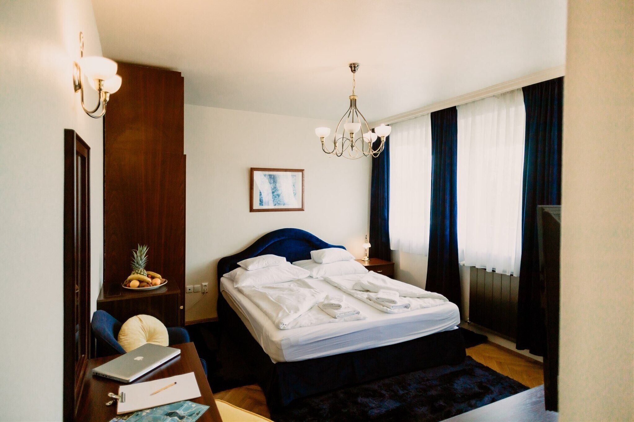 A hotel room with a neatly made bed, a desk with a laptop and papers, a fruit bowl, a painting on the wall, and a chandelier.