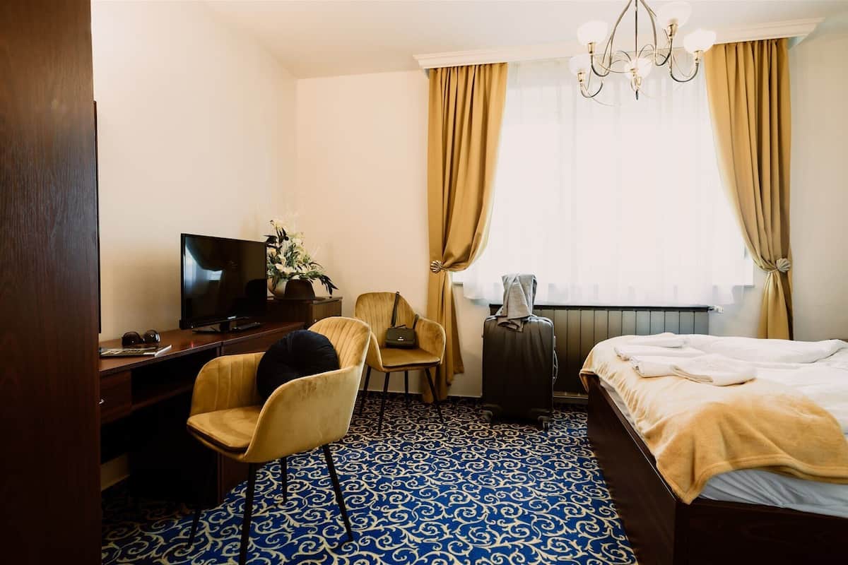 A hotel room with a bed, a desk with a television, a chair, a suitcase, and a chandelier.