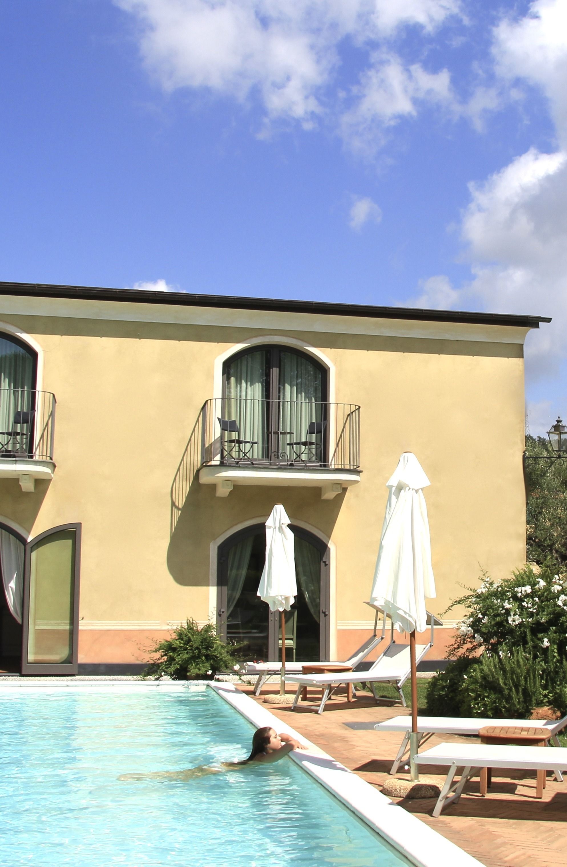 Una piscina con lettini e ombrelloni, un edificio con balconi e ampie finestre, e un cielo azzurro limpido.