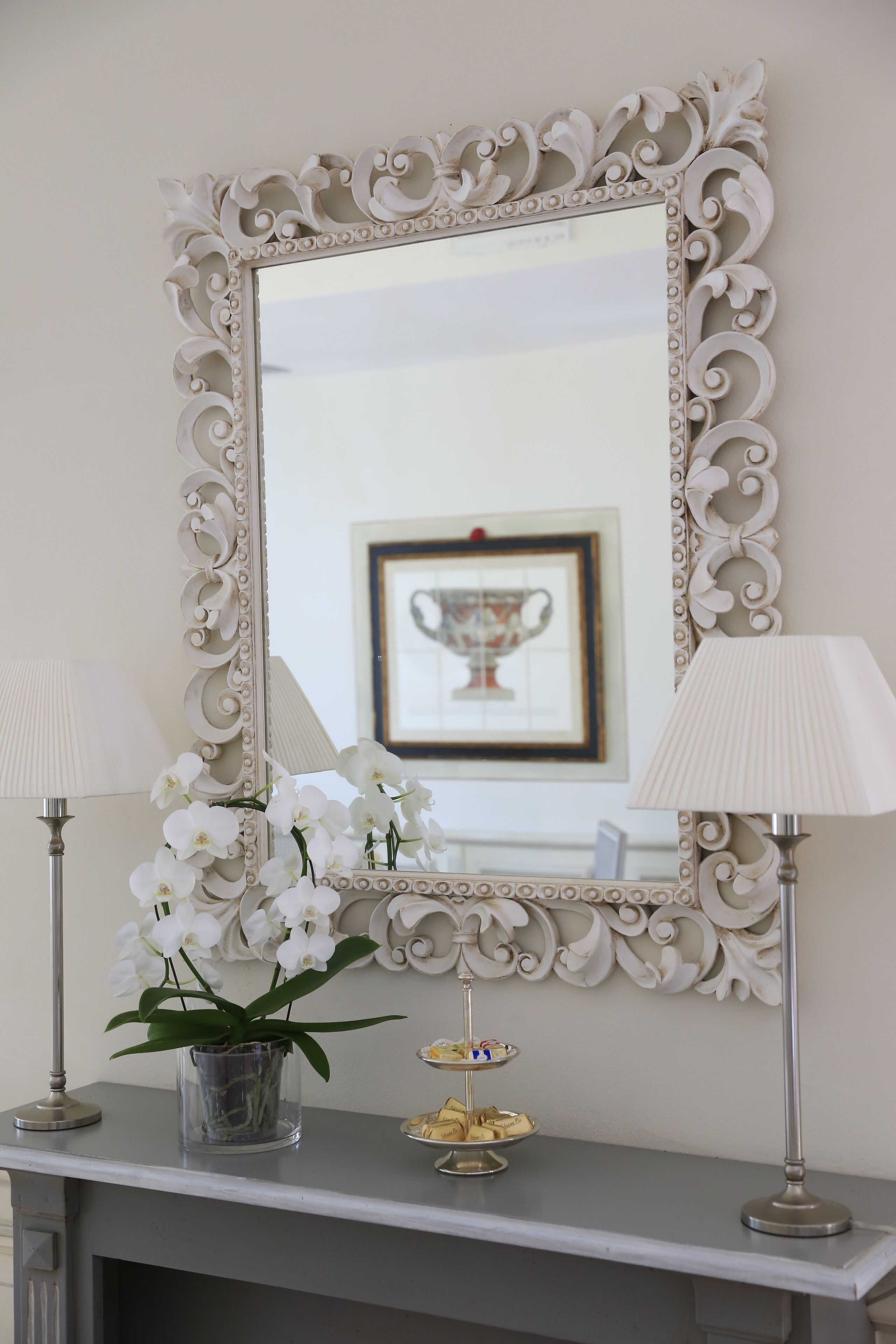 A mirror with an ornate frame, a vase with white flowers, and a tray with cookies on a mantelpiece.