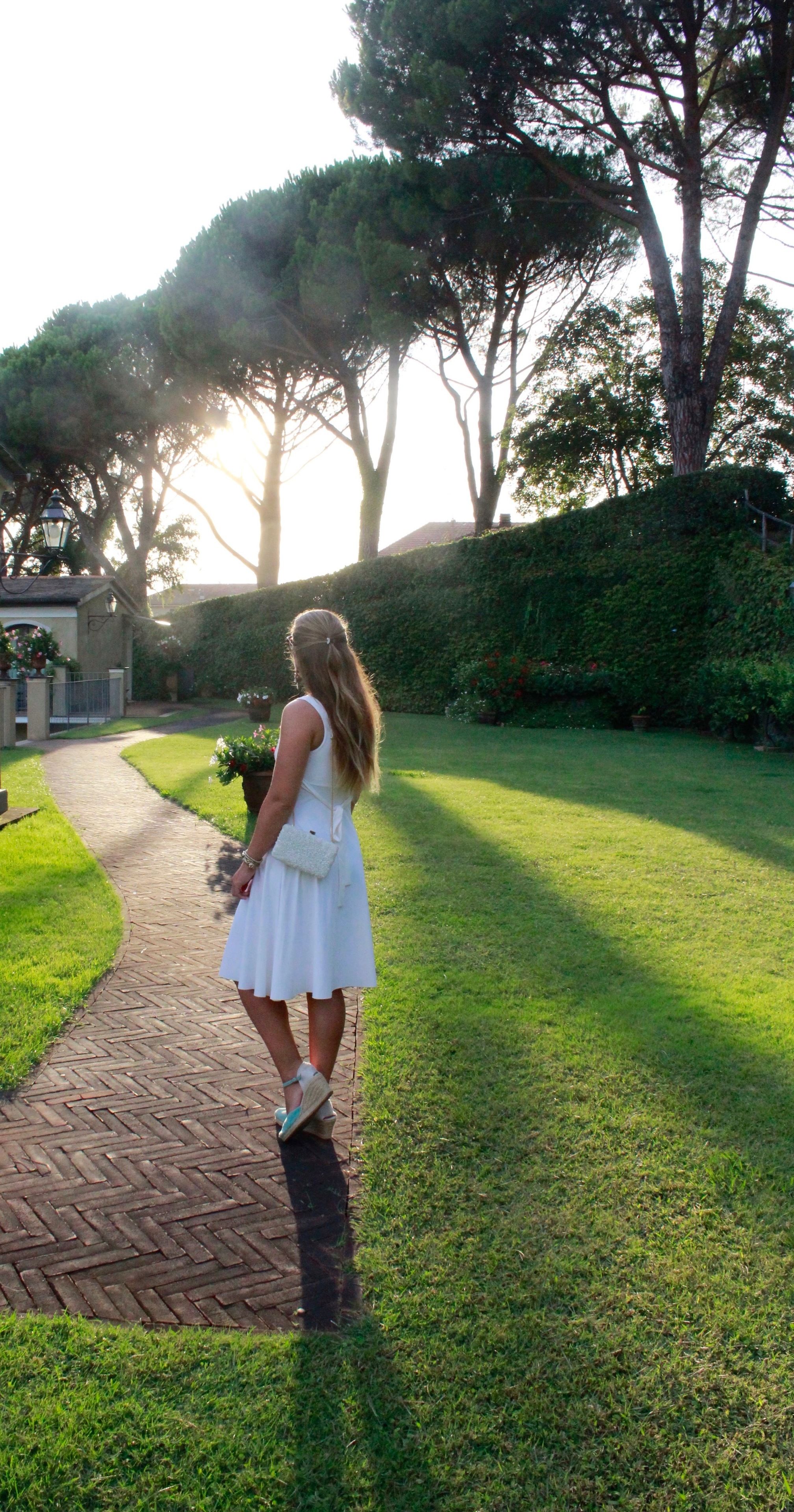 A woman in a white dress walking on a brick pathway in a garden with trees and a lamp post.