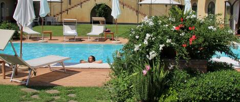 Un área de piscina con sillones de descanso y sombrillas, rodeada de vegetación y un camino de piedra.