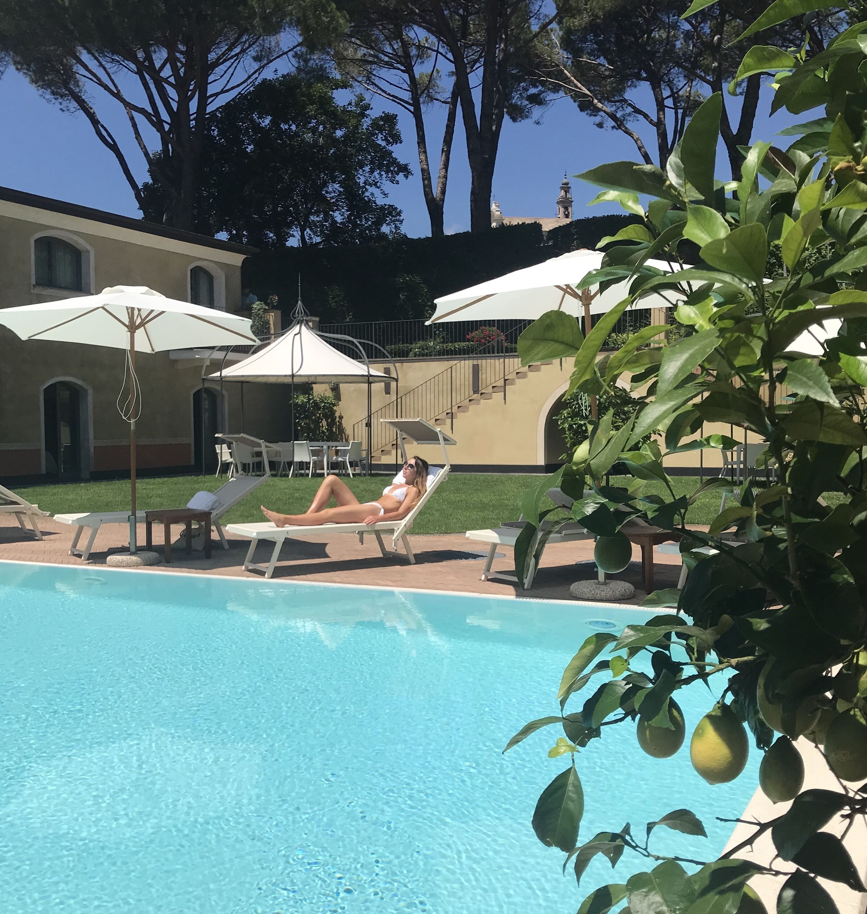 Zona de piscina con sillas de descanso y sombrillas, rodeada de vegetación y un edificio con una torre de reloj al fondo.
