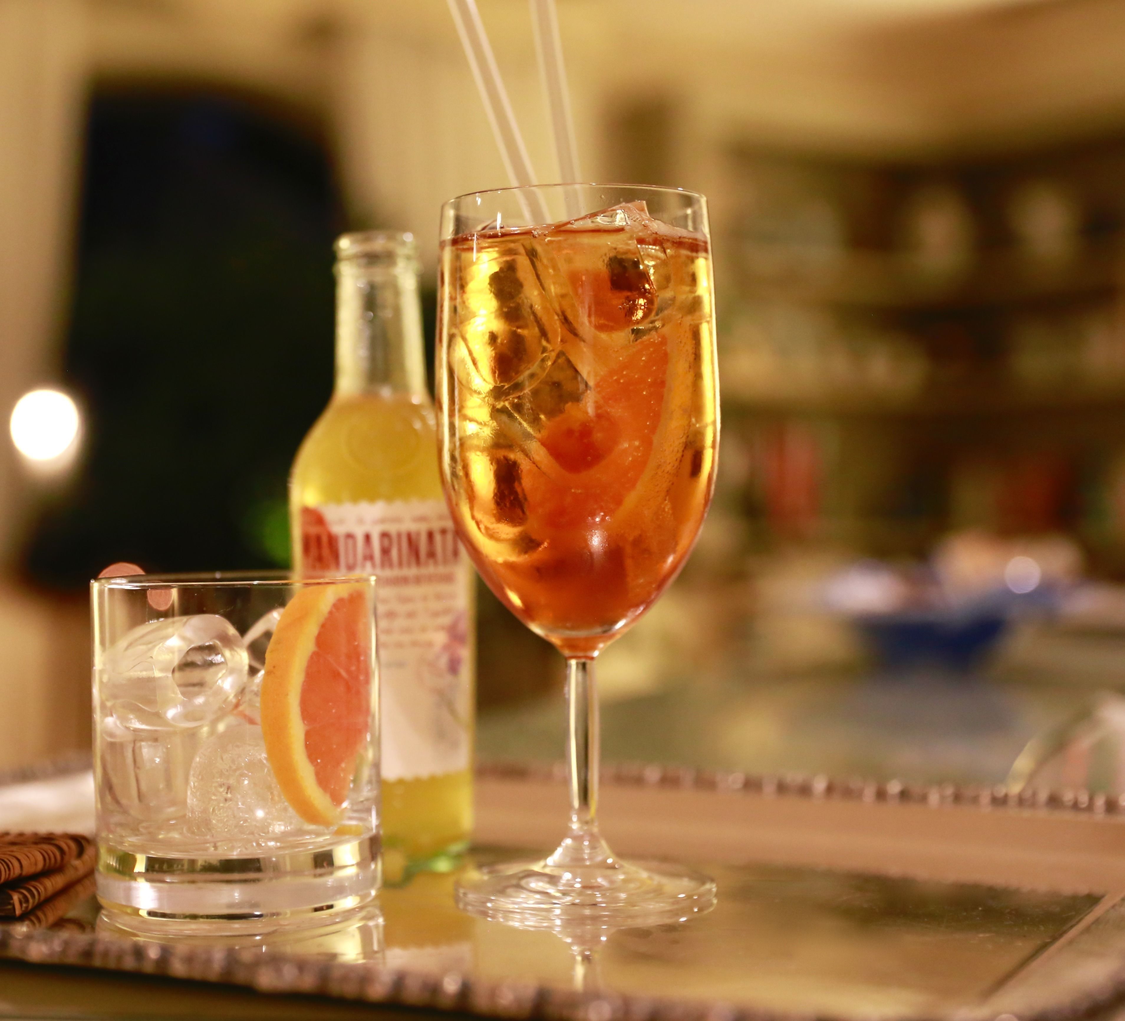 A bar setting with a glass of amber-colored drink, ice cubes, and a slice of orange. A bottle labeled "MANDARIN" is visible in the background.