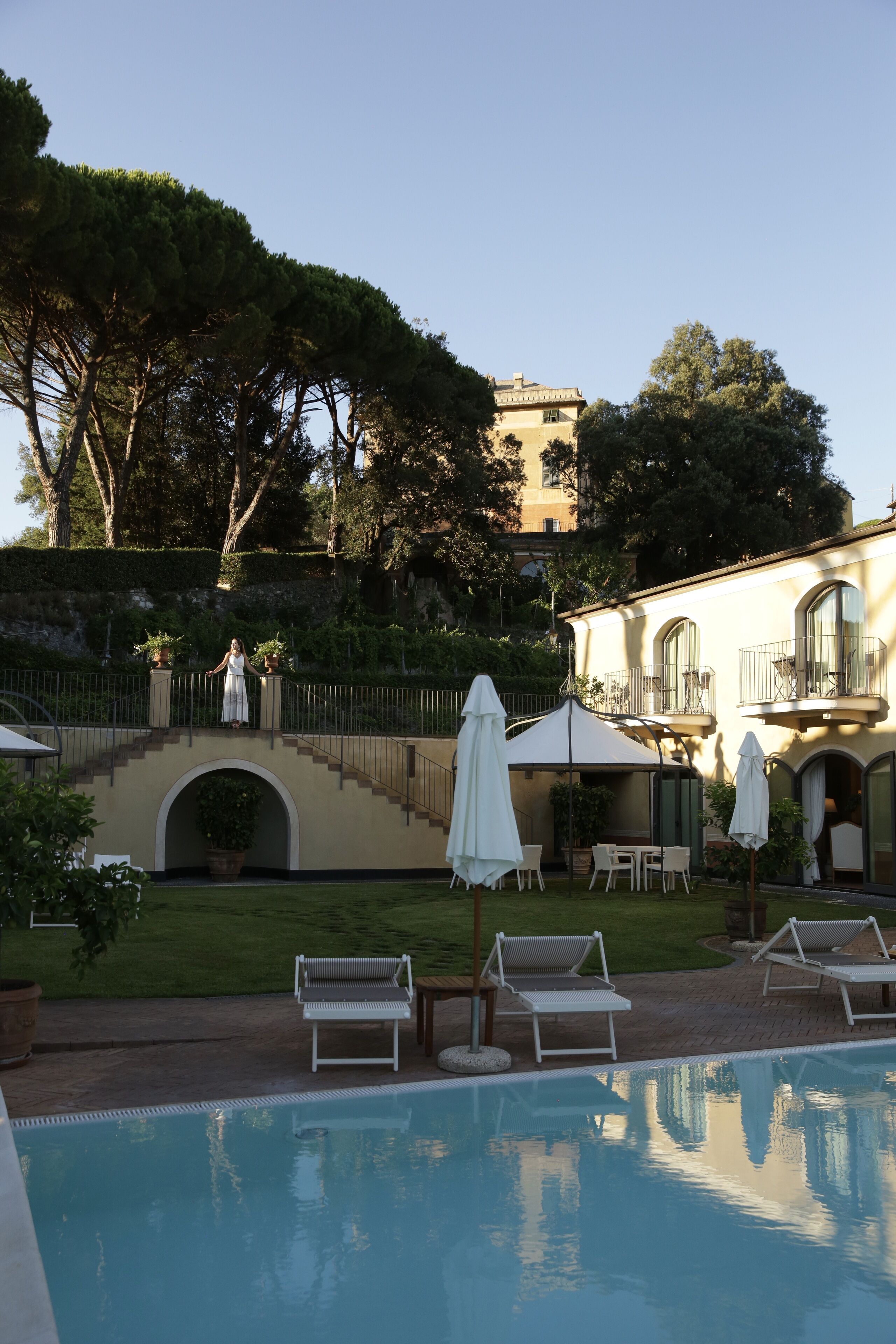 A pool area with lounge chairs and umbrellas, a bridge, and a building with a garden and trees in the background.