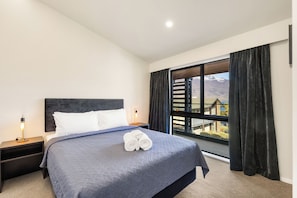 A hotel room with a bed, a nightstand, a lamp, a window with curtains, and a view of mountains. - Queenstown Motel Apartments (Queenstown)