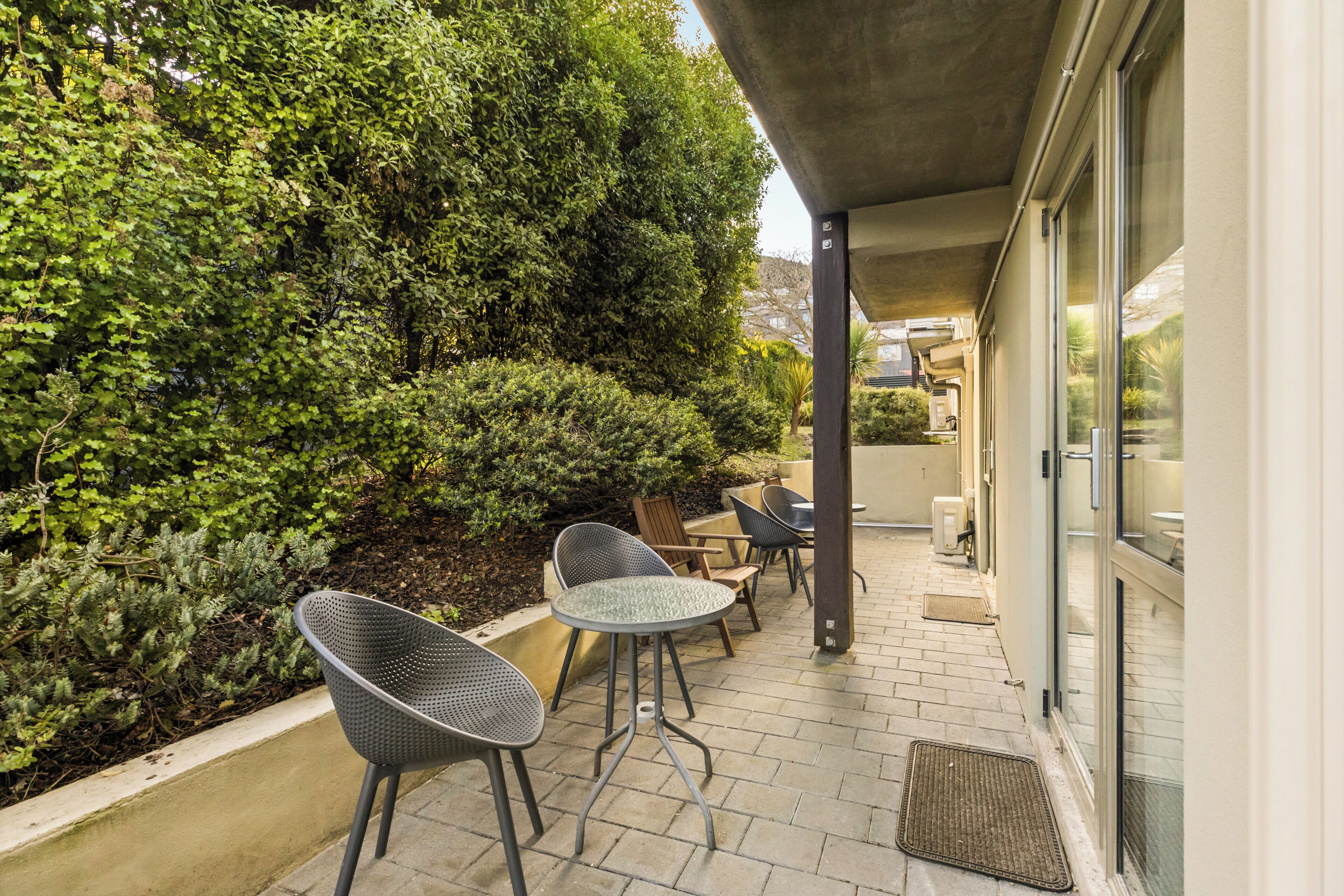 An outdoor patio with chairs and tables, surrounded by greenery and a building with large windows.