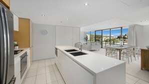 A modern kitchen with a central island, a dining area with a table and chairs, and a large window offering a view of the city. - Ambience on Burleigh Beach (Burleigh Heads)