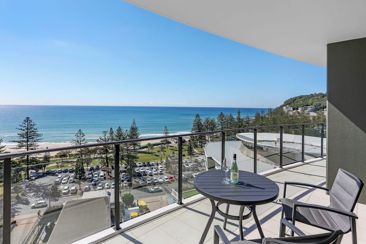 Uma varanda com mesa e cadeiras, com vista para a praia e um estacionamento.