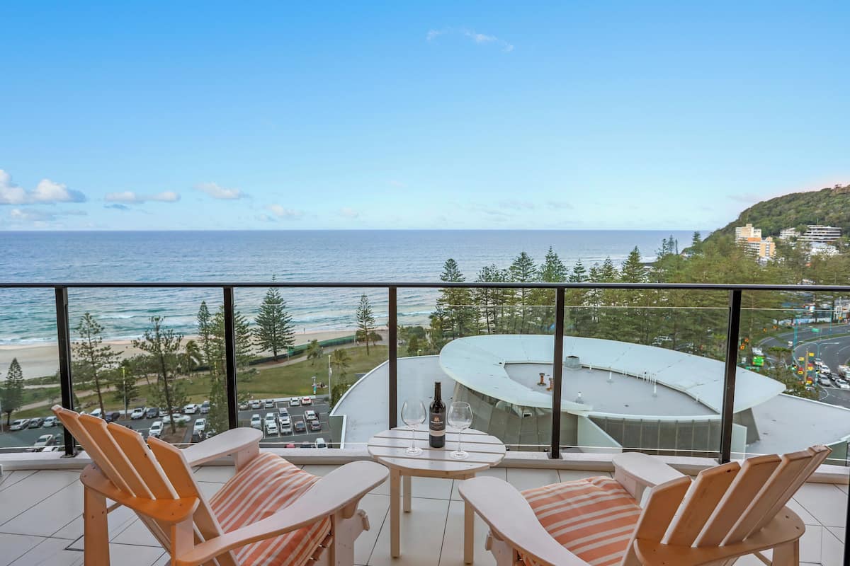 Varanda com duas cadeiras e uma mesa, com vista para uma praia e um edifício redondo.