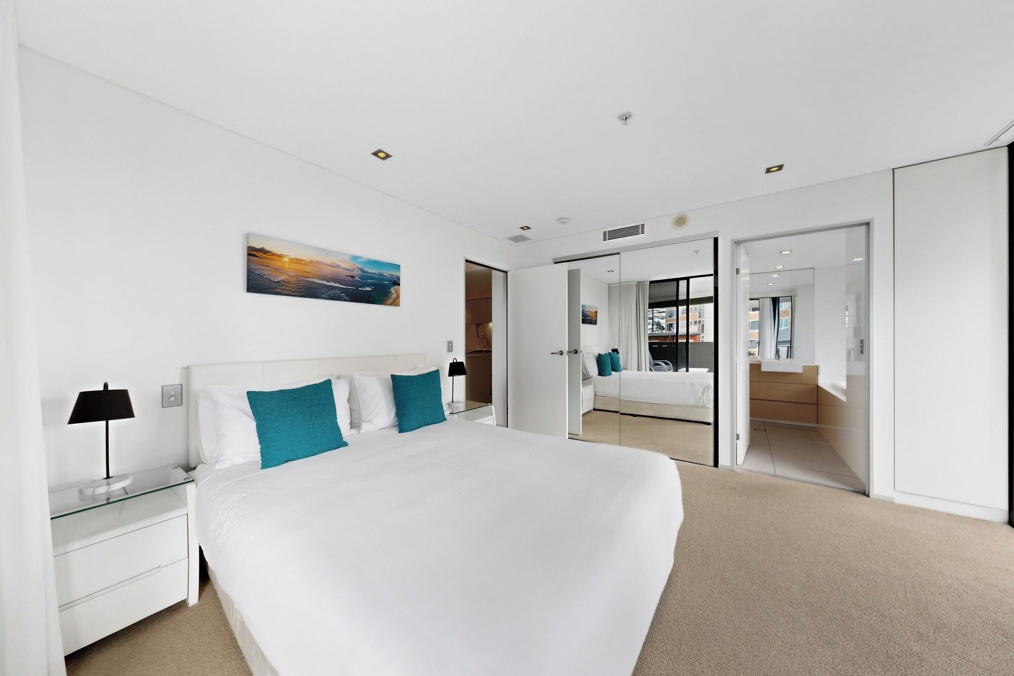 A modern hotel room with a large bed, a nightstand, a lamp, a painting, and a view into the bathroom.