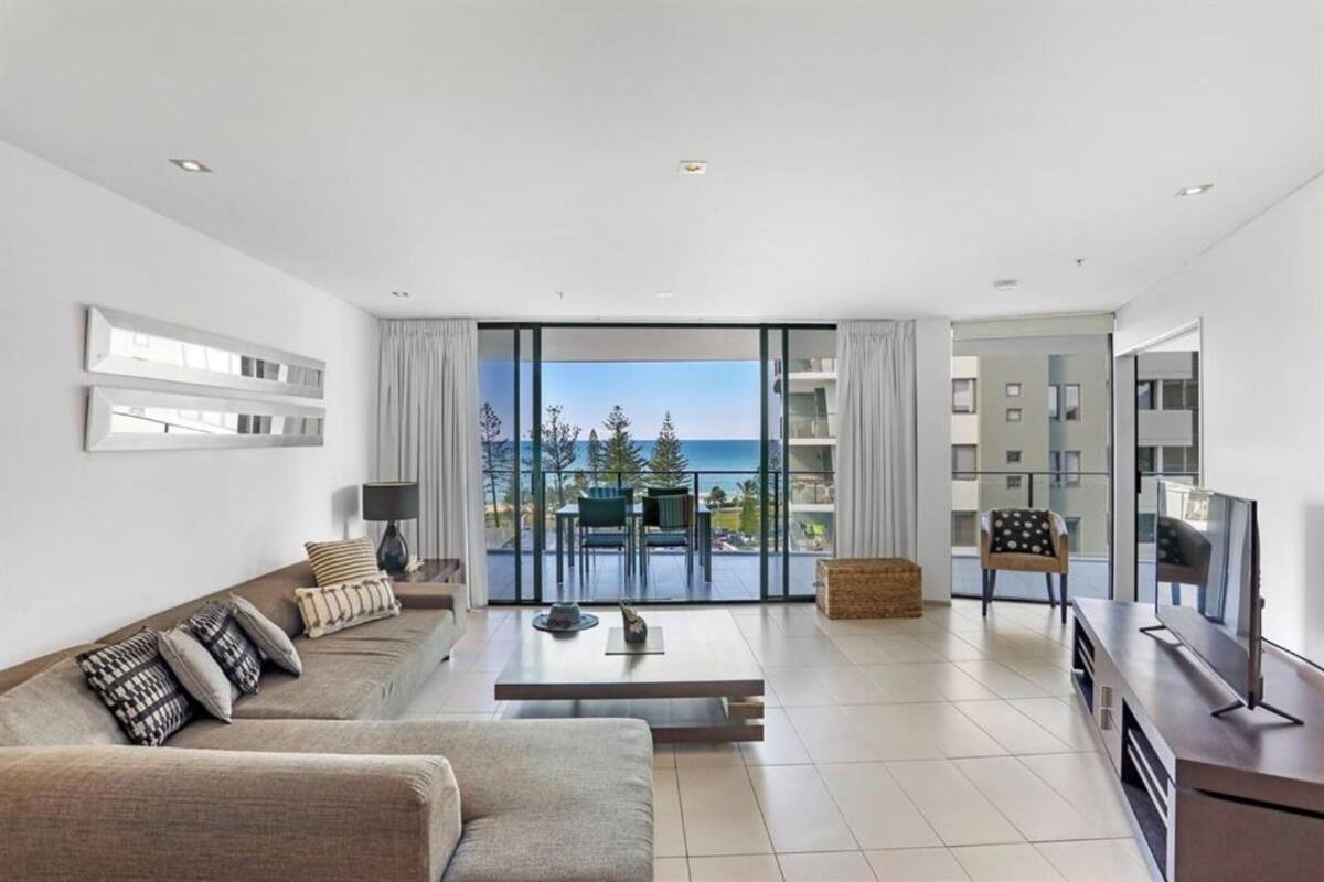 Uma sala de estar moderna com sofá, mesa de centro e televisão, janelas amplas com vista para o mar e uma varanda com mobiliário para exterior.