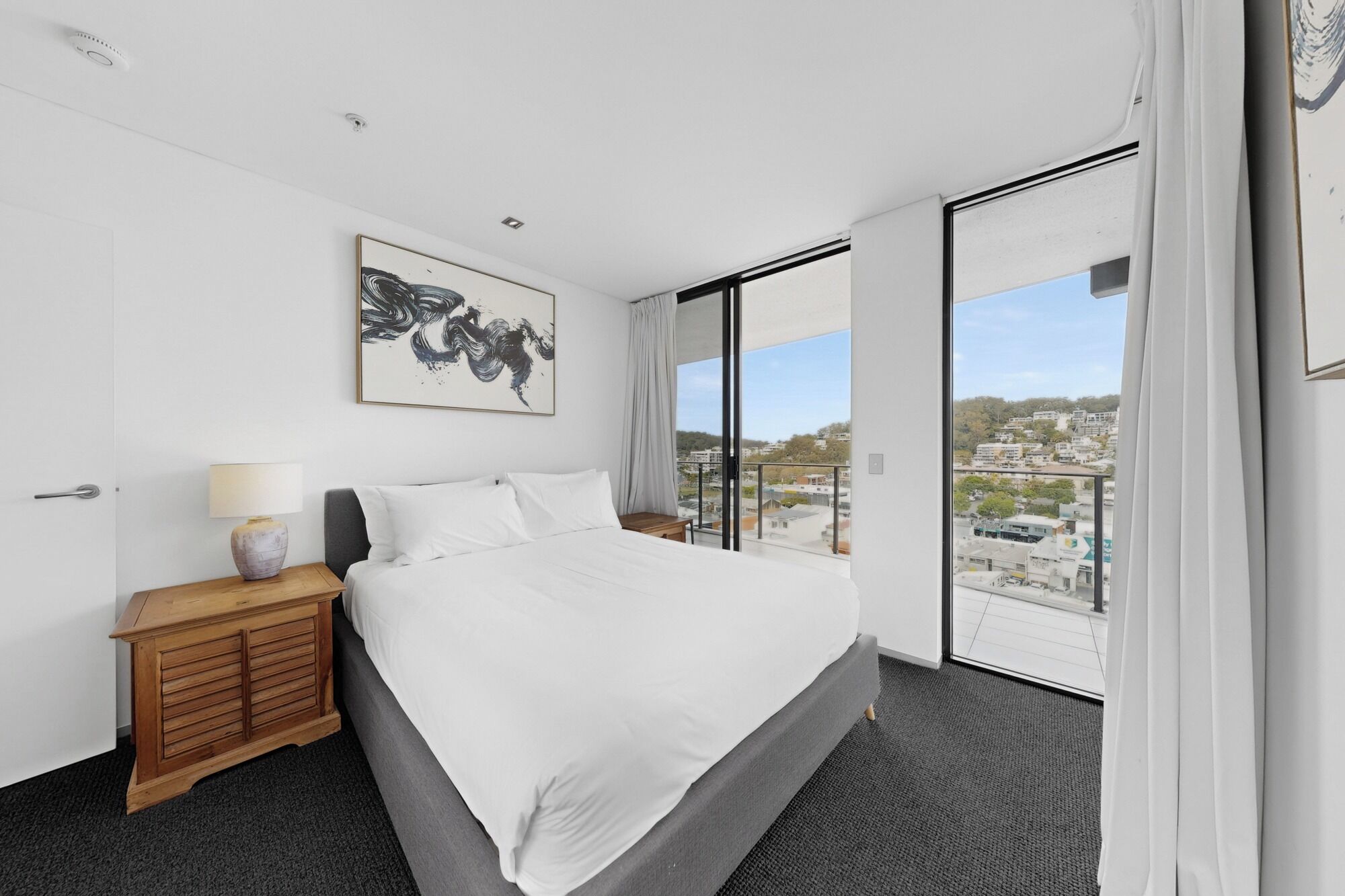 Habitación de hotel con cama, mesita de noche, lámpara, un cuadro y un ventanal con vistas a la ciudad.