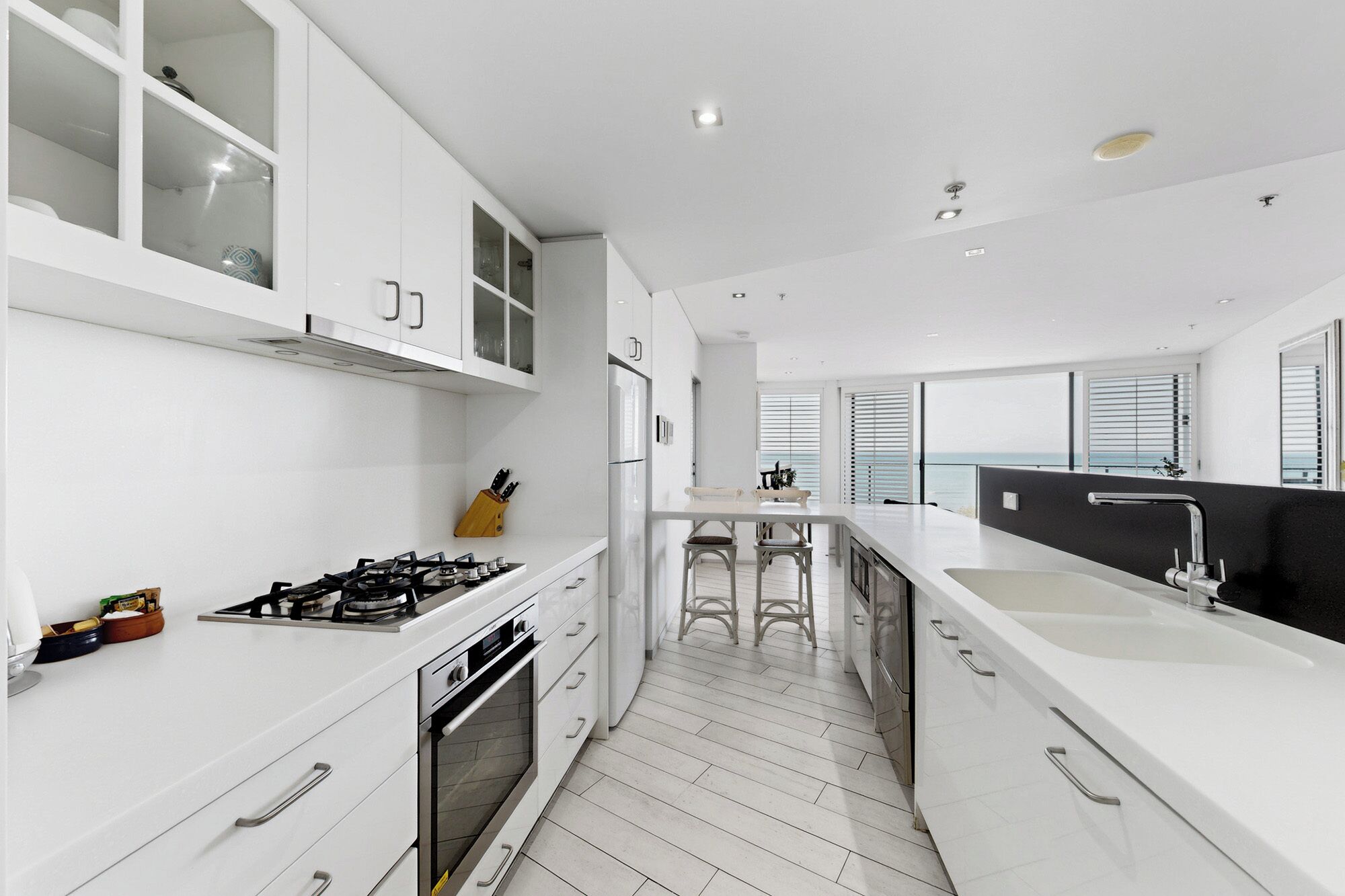 A modern kitchen with white cabinets, a gas stove, an oven, and a large window overlooking the sea.