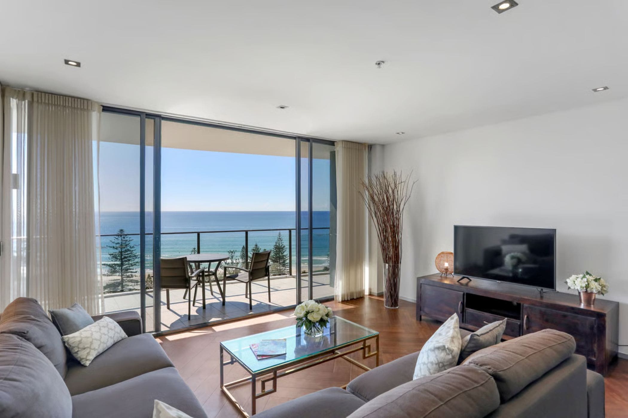 A modern living room with a large flat-screen TV, a sofa, a coffee table, and a balcony with a view of the ocean.
