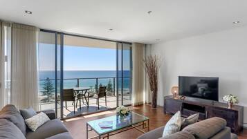 A modern living room with a large flat-screen TV, a sofa, a coffee table, and a balcony with a view of the ocean.