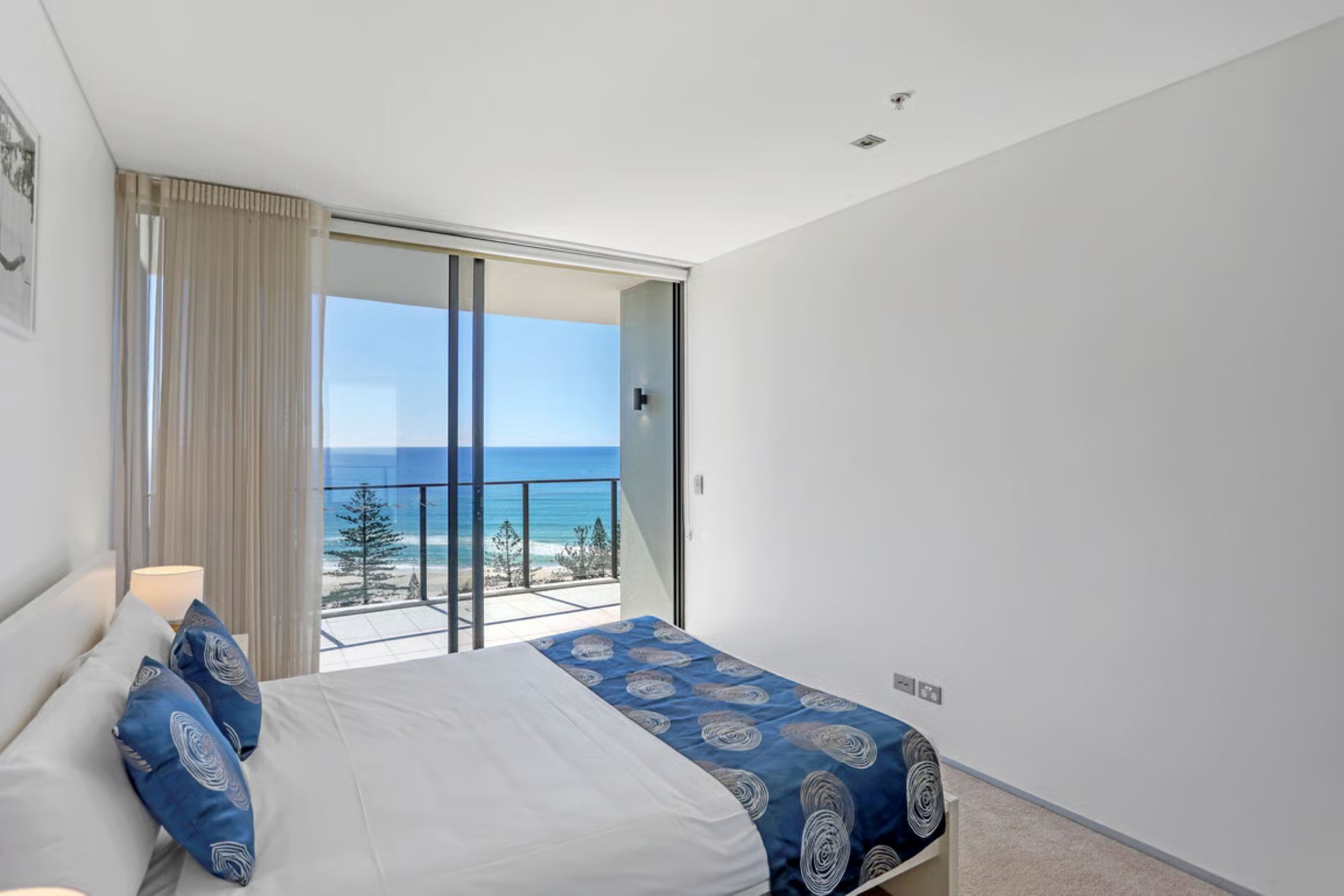 Un dormitorio con cama, mesita de noche, lámpara, vista al océano y balcón con puerta de vidrio.