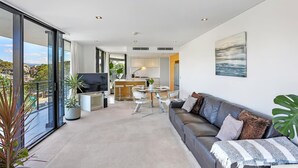 A modern living area with a sofa, TV, dining table, and kitchen in the background. - Ambience on Burleigh Beach (Burleigh Heads)