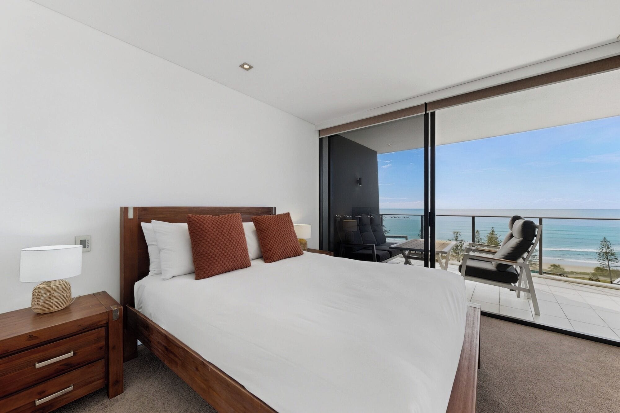 A bedroom with a large bed, a nightstand, a lamp, and a view of the ocean through a balcony.