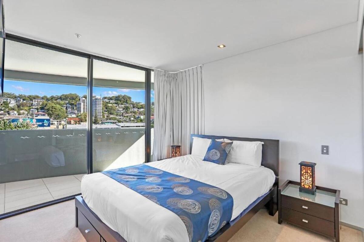 Quarto de hotel moderno com uma cama grande, divisória de vidro, mesa de cabeceira e vista da cidade.
