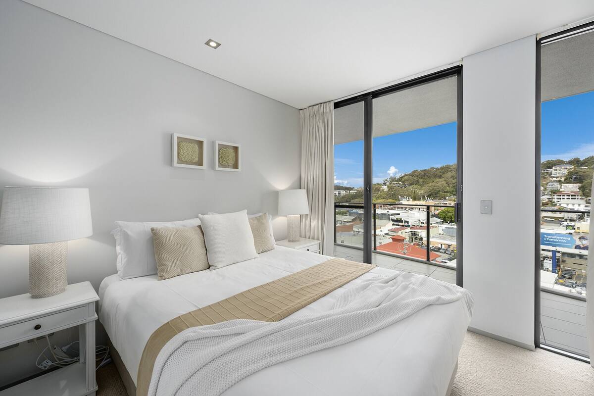 Quarto de hotel moderno com uma cama grande, duas mesas de cabeceira com luminárias, vista para a cidade através de uma porta de vidro deslizante e duas obras de arte emolduradas na parede.