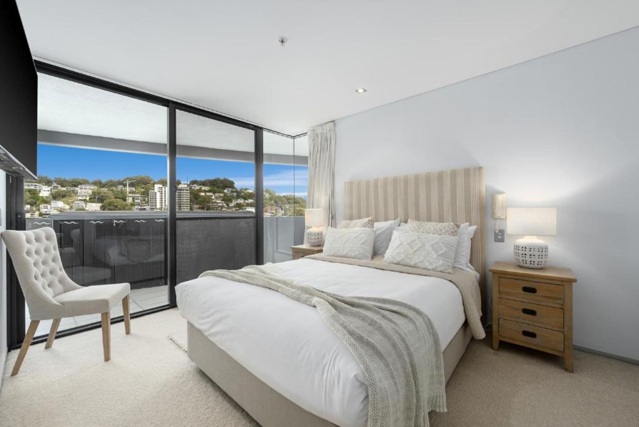 A bedroom with a large bed, a chair, a nightstand, and a view of the city through a balcony.