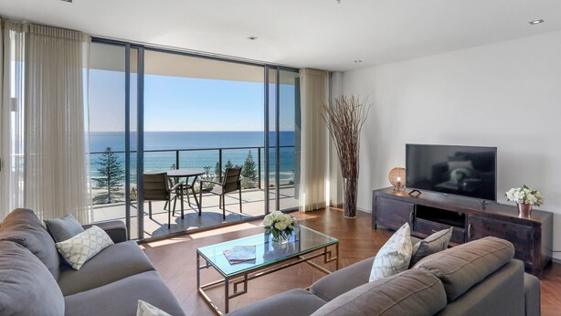 A modern living room with a large TV, comfortable sofas, a glass coffee table, and a balcony with a view of the ocean. - Ambience on Burleigh Beach (Burleigh Heads)