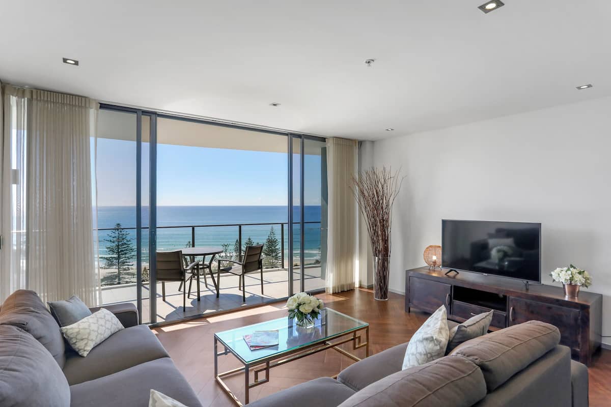 Uma sala de estar moderna com uma televisão grande, sofás confortáveis, uma mesa de centro de vidro e uma varanda com vista para o oceano.