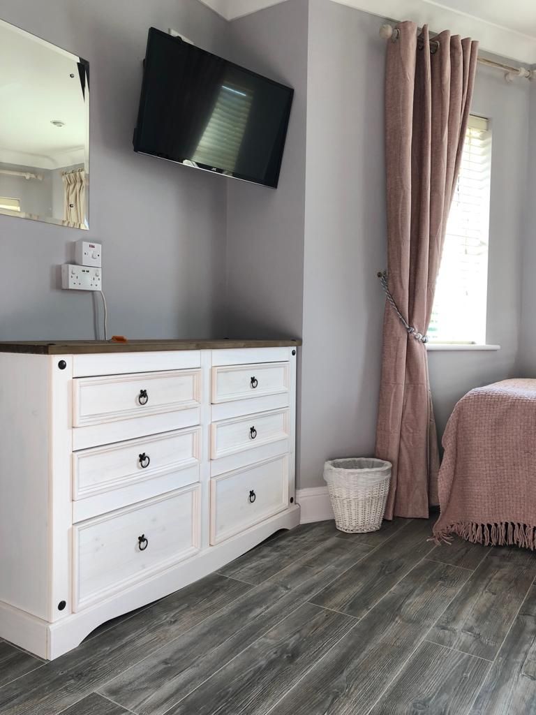 A bedroom with a white dresser, a television mounted on the wall, a window with curtains, and a bed with a pink blanket.