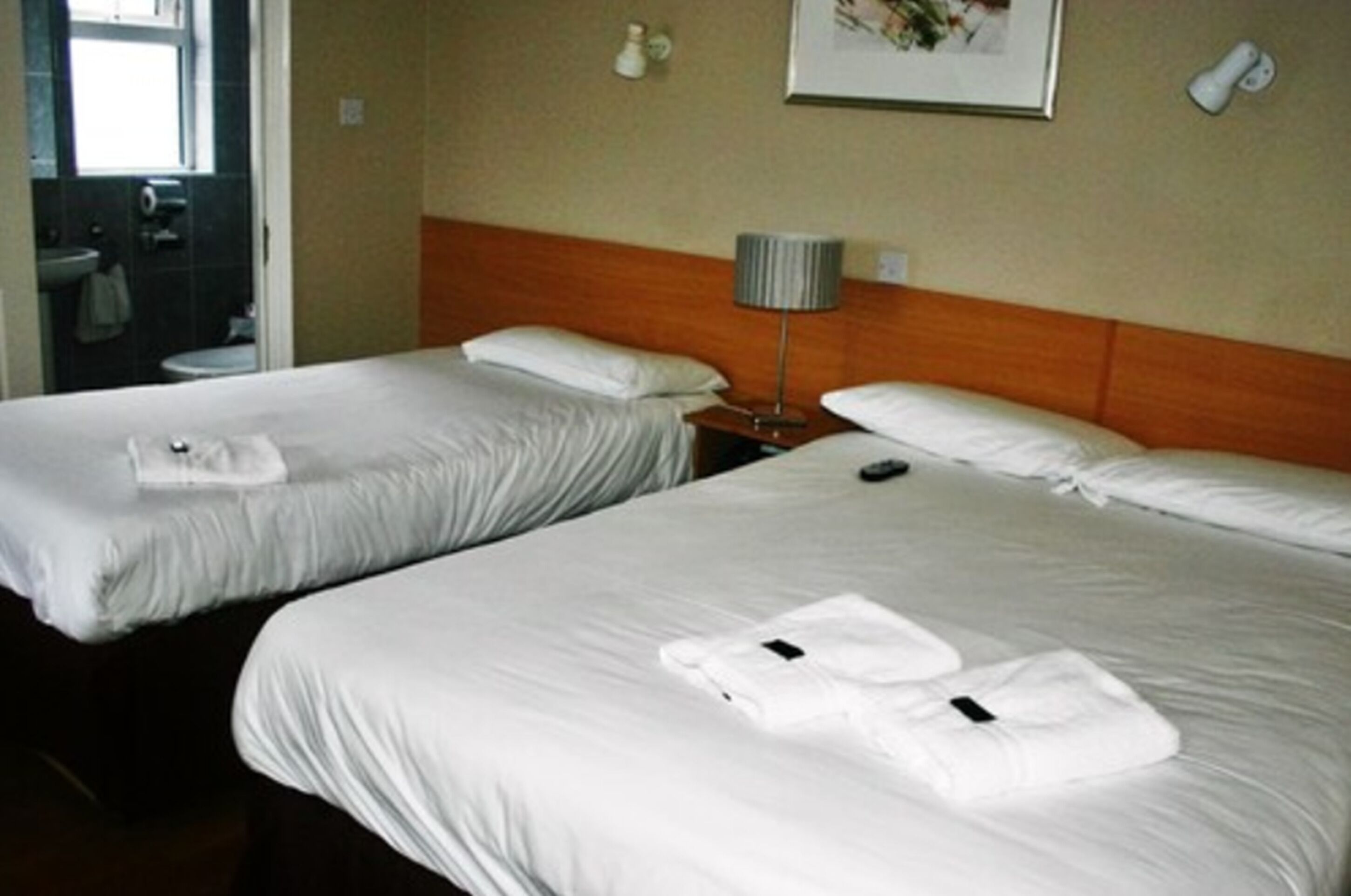 Habitación de hotel con dos camas individuales, una mesita de noche con lámpara, un cuadro en la pared y un baño visible a través de una puerta abierta.