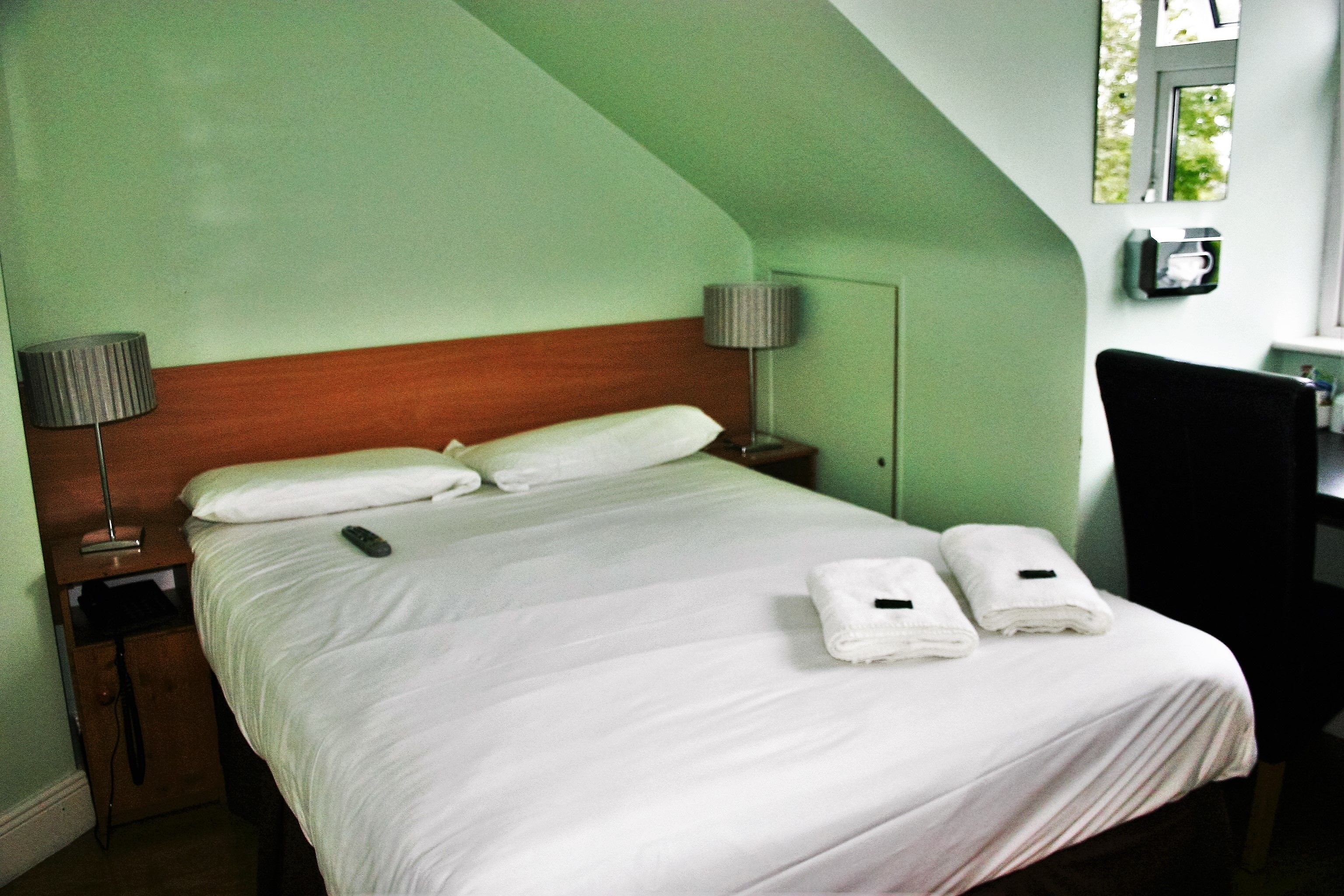 A neatly made bed with white linens and two folded towels, a wooden headboard, a nightstand with a lamp, and a desk with a chair.