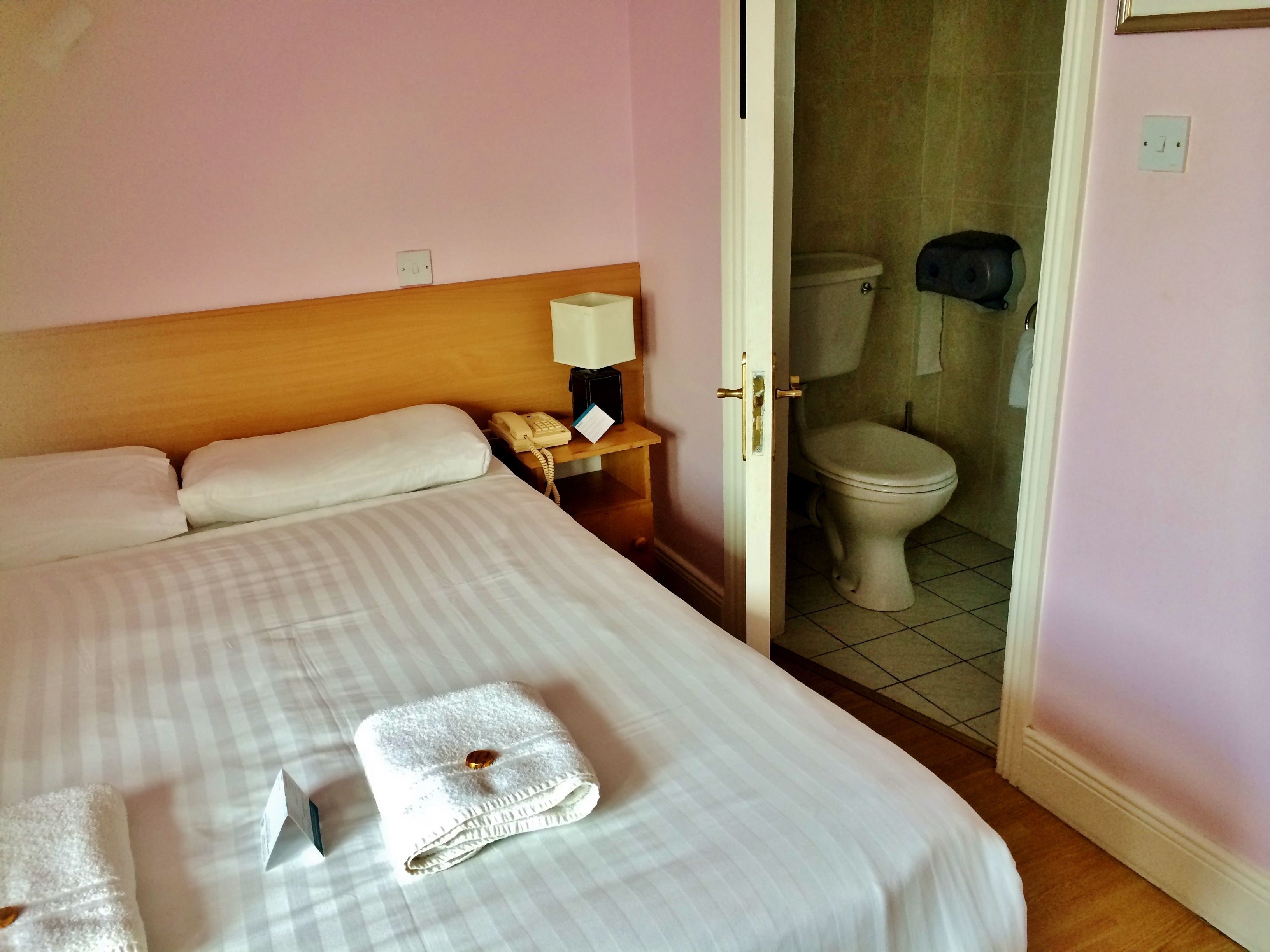 Habitación de hotel con cama, teléfono en la mesita de noche, una lámpara y un inodoro visible a través de una puerta abierta.