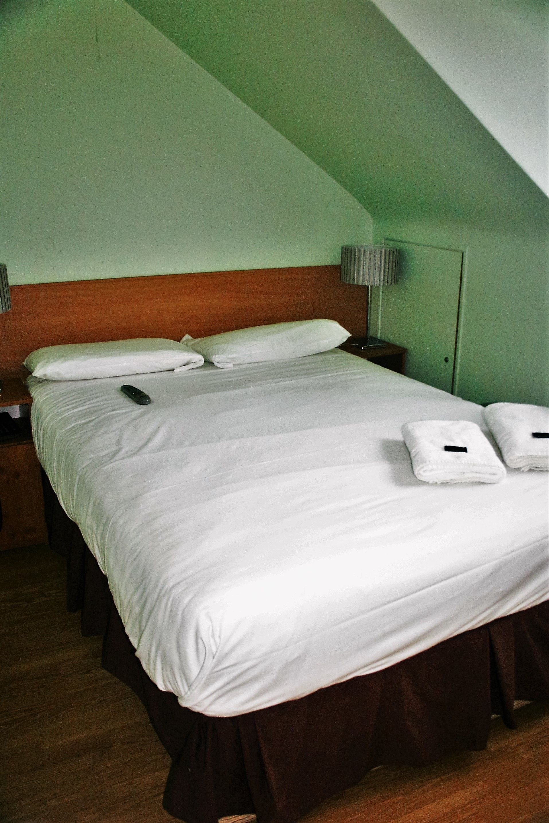 Una cama bien hecha con sábanas blancas, un control remoto y toallas dobladas sobre un cabecero de madera en una habitación con paredes verdes y techo inclinado.