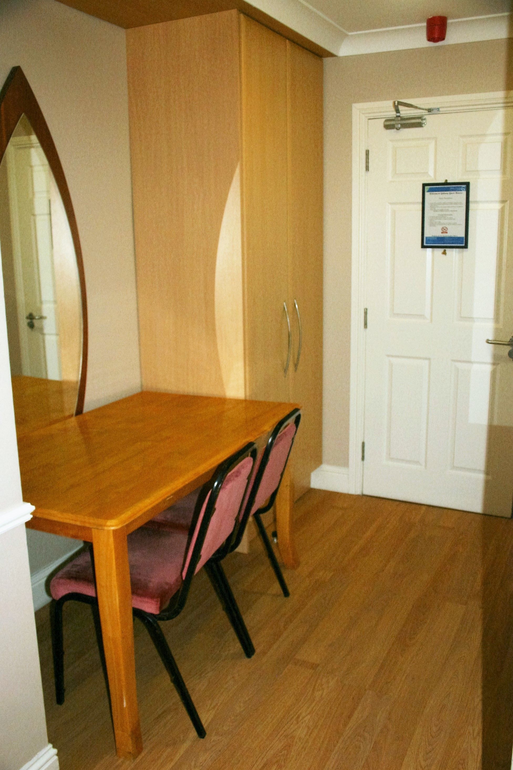 Une table à manger en bois avec des chaises, une porte blanche avec un avis affiché, et un miroir au mur.