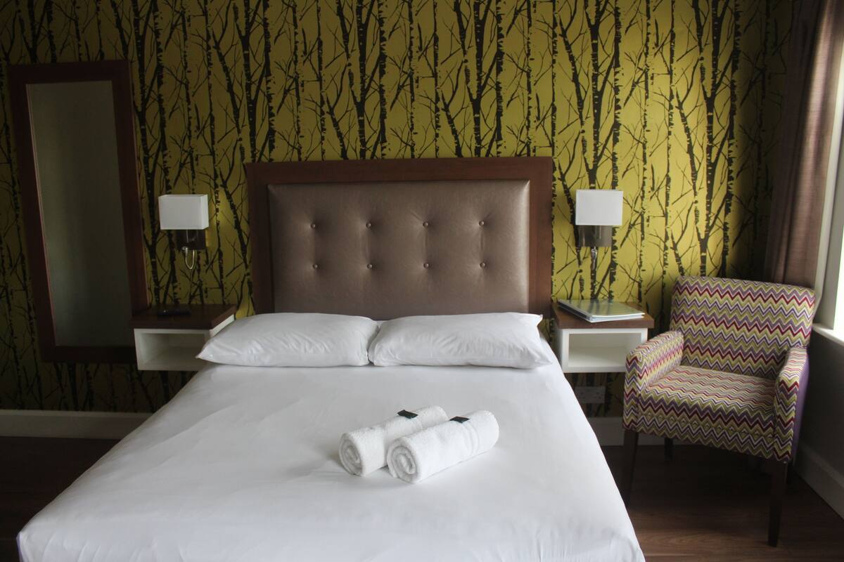 A hotel room with a large bed, two white pillows, two rolled towels, a wooden headboard, two bedside tables with lamps, a patterned chair, and a mirror on the wall.