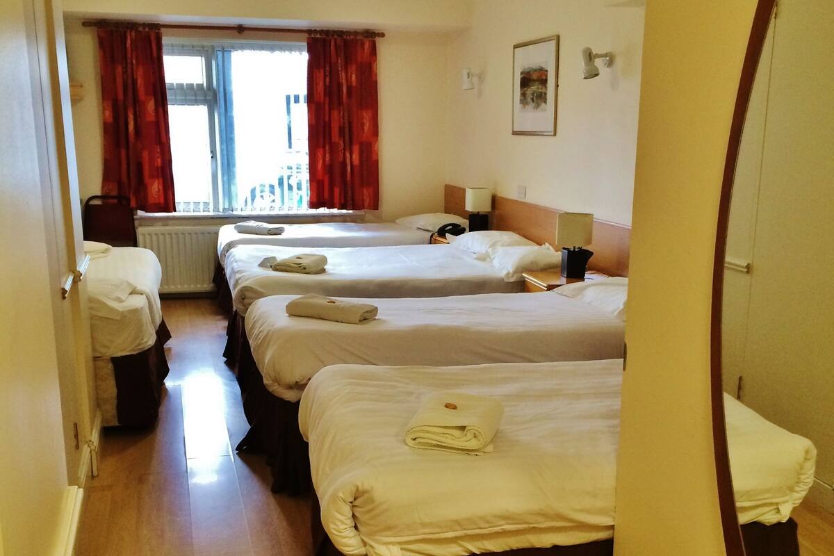 A hotel room with three beds, a radiator, a window with red curtains, a bedside table with a lamp, and a mirror on the wall.