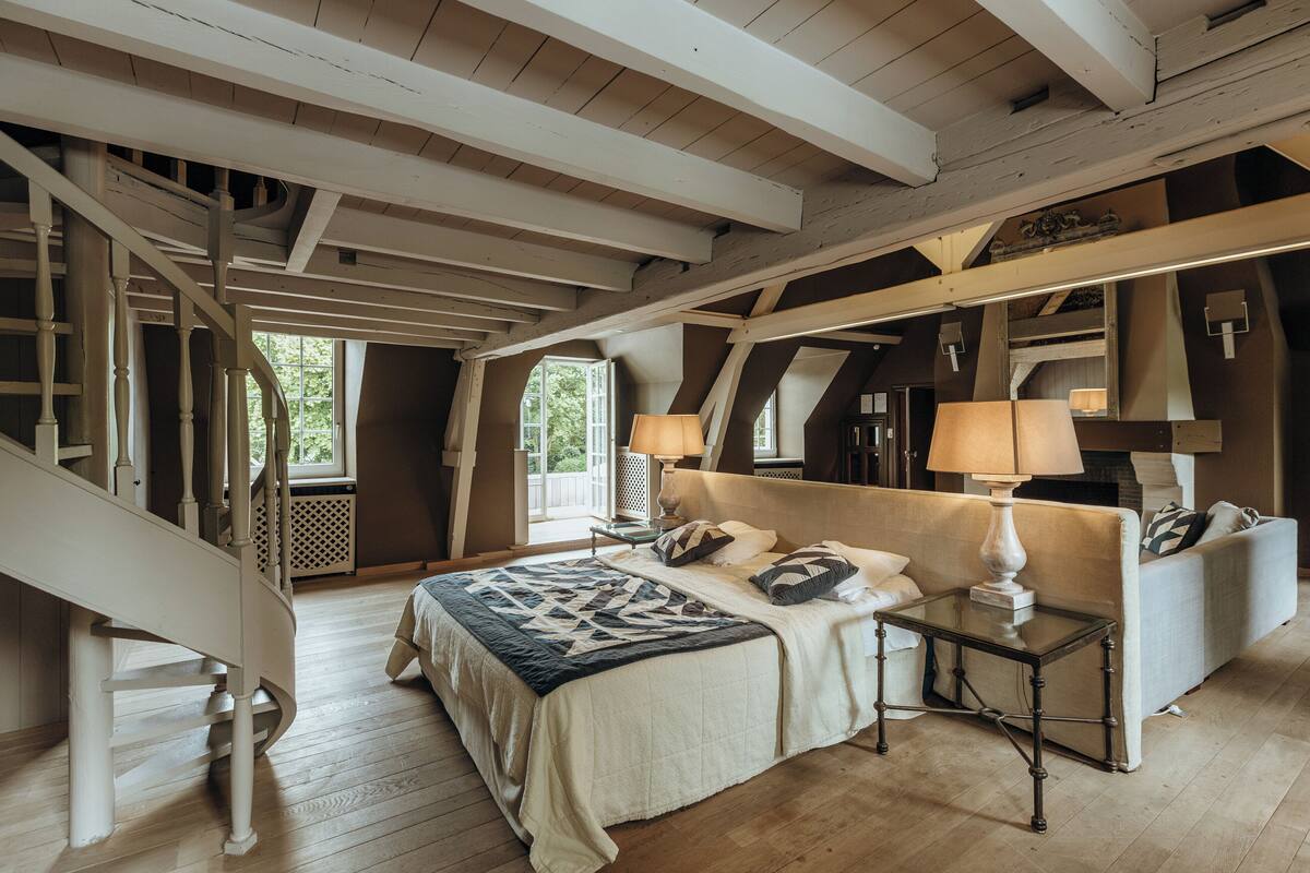 A bedroom with a large bed, a nightstand, a lamp, a sofa, and a staircase.
