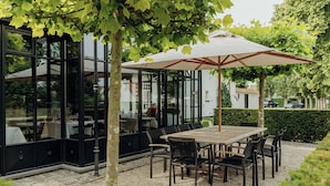Un área de comedor al aire libre con una mesa y sillas de madera, un paraguas grande y paredes de vidrio.