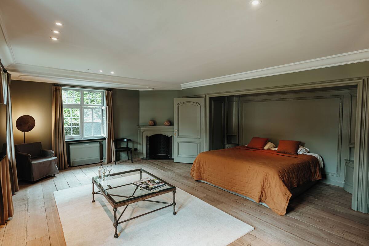 A bedroom with a large bed, a fireplace, a sitting area with a chair, and a table with magazines.