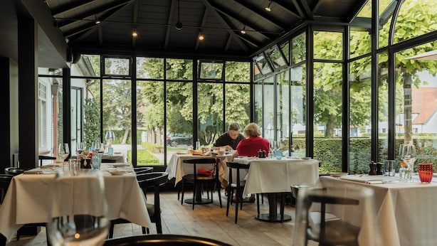 Un comedor interior con mesas dispuestas para dos, ventanas amplias y vistas al exterior.
