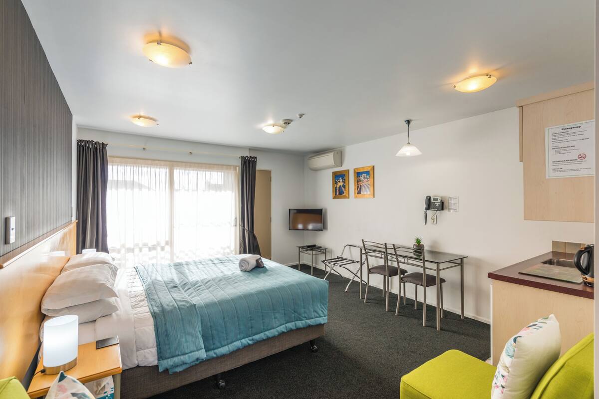 A hotel room with a bed, bedside table, lamp, TV, desk, chair, and a small kitchenette.