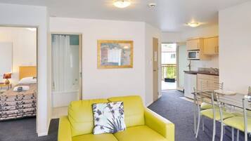 A bright yellow sofa with a floral pillow in a room with a dining area and a kitchen visible through an open door.