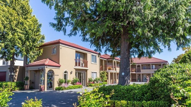 Ein mehrgeschossiges Hotel mit rotem Ziegeldach, einem begrünten Vorgarten und einem großen Baum, der Schatten spendet.