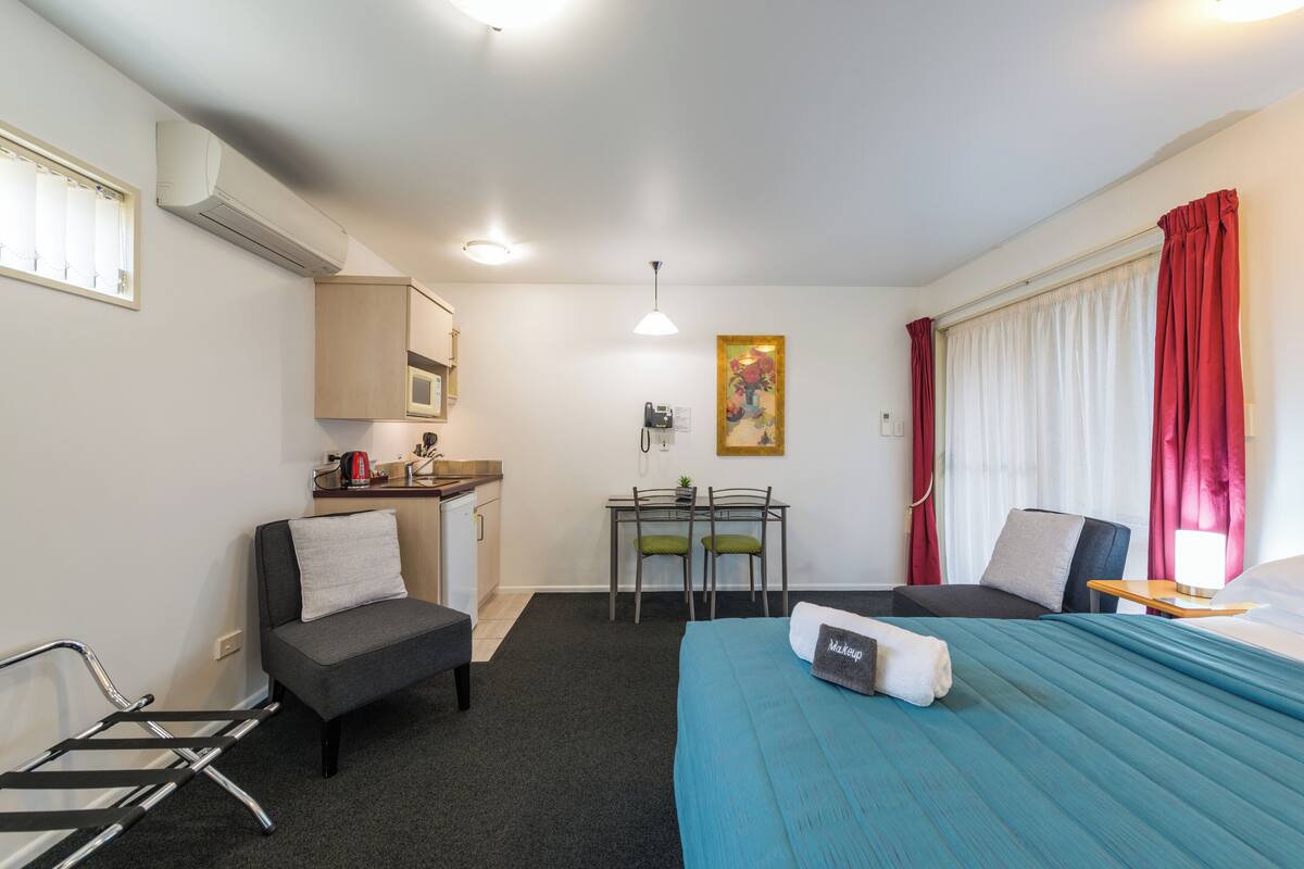 A compact hotel room with a bed, a desk with a chair, a kitchenette, and a window with curtains.