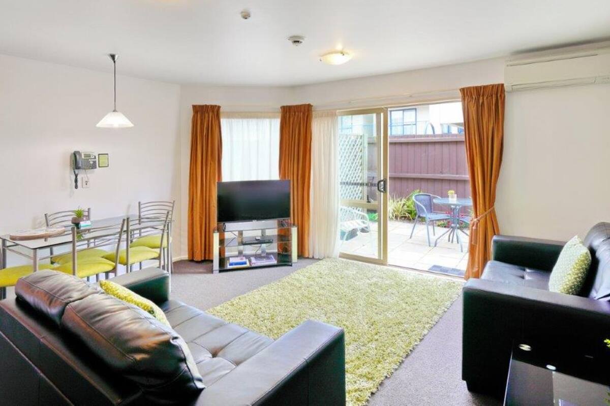 A modern living room with a sofa, a coffee table, and a TV on a glass stand. There is a sliding glass door leading to an outdoor area with a table and chairs.