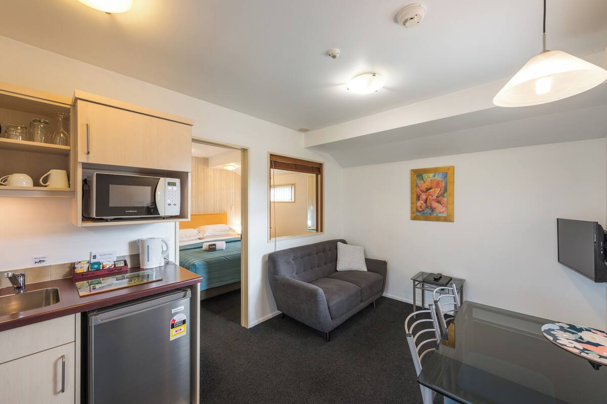 A compact hotel room with a kitchenette, a sofa, a dining table, and a TV.