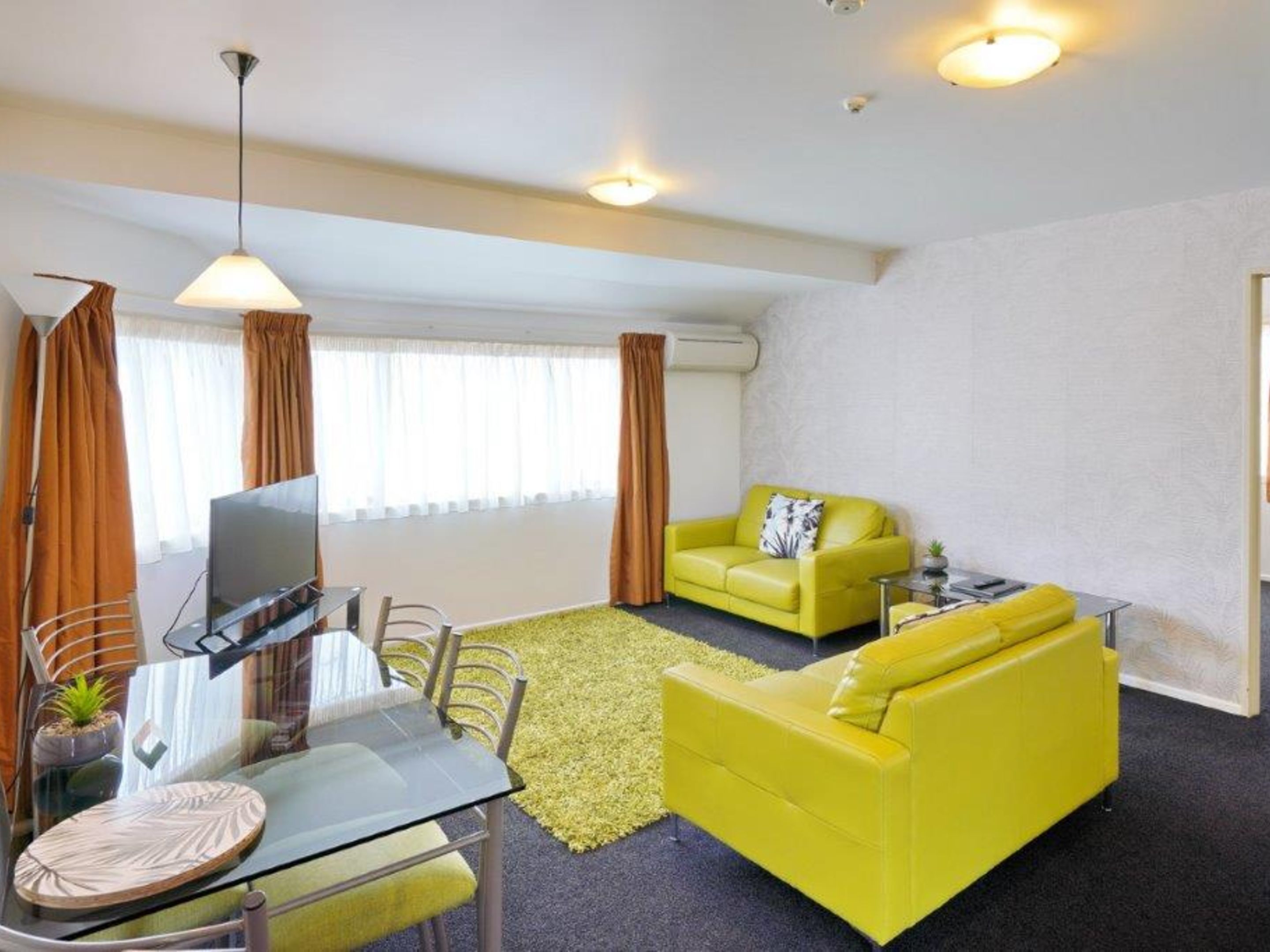 A modern living room with a glass dining table, yellow sofas, and a large window with curtains.